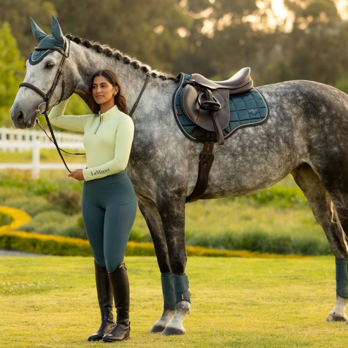 Woman in equestrian attire standing next to a grey horse in a field