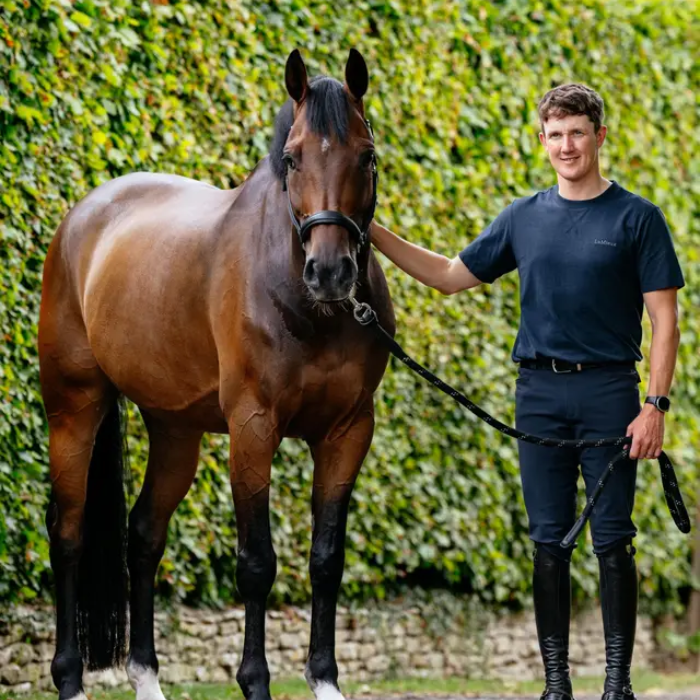 Man wears a green t-shirt and navy breeches holding a horse against a leafy background