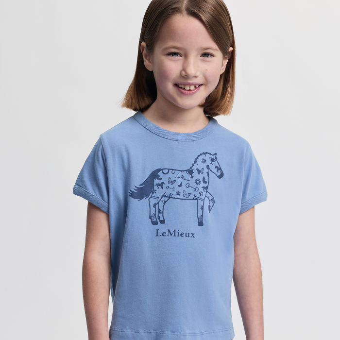 Young girl wearing a blue t-shirt with a horse design and 'LeMieux' branding on a light grey background.