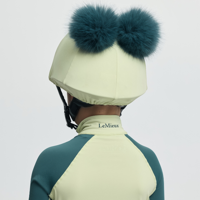 Person wearing a green and yellow equestrian helmet with teal fur pom-poms on a light grey background.