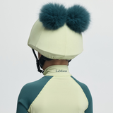 Person wearing a green and yellow equestrian helmet with teal fur pom-poms on a light grey background.