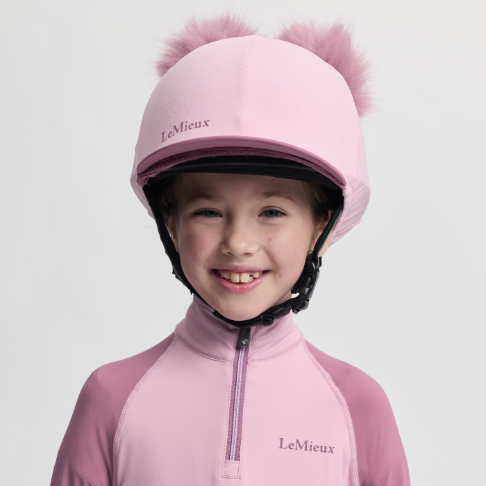 Child wearing a pink equestrian helmet with pom-poms and matching outfit on a light grey background