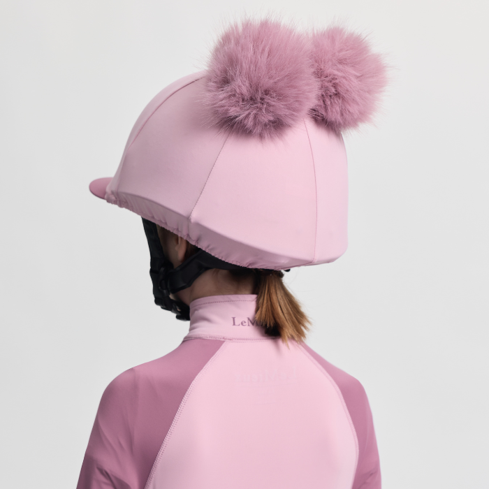Person wearing a pink equestrian helmet with a large fur pom-pom on a light grey background