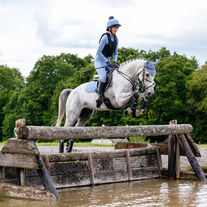 Equestrian jumping over a wooden obstacle into water with trees in the background