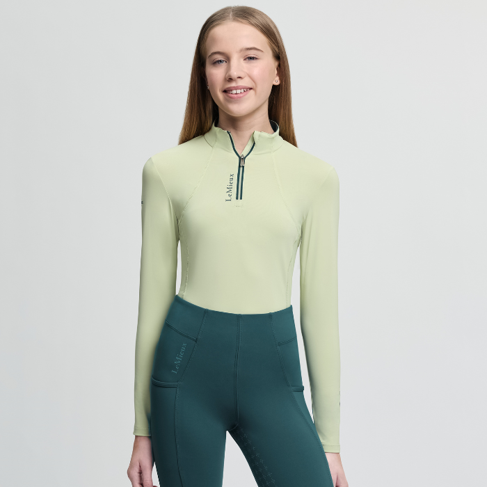 Person wearing a light green long-sleeve top and teal riding pants on a grey background