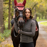 LeMieux AW26 Maddie Hybrid Jacket Varient Cocoa. A woman stands beside a dark brown horse on a wooded path, wearing a fitted ribbed equestrian jacket over a red top. The horse wears a red fly veil and black bridle.