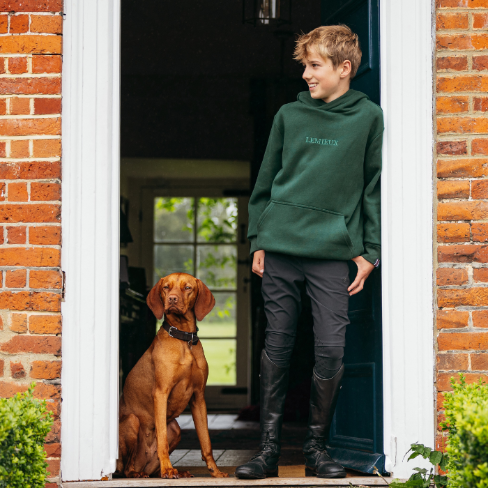 Person wearing a green hoodie standing next to a dog at a door.