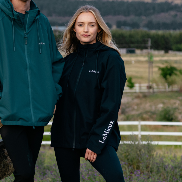 Woman wears a navy waterproof jacket with LeMieux branding in a blurred outdoor setting