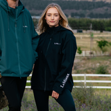 Woman wears a navy waterproof jacket with LeMieux branding in a blurred outdoor setting