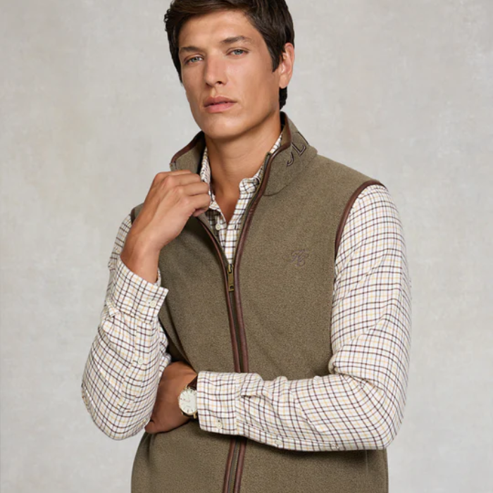 Man wears a light khaki fleece gilet over a plaid shirt on a light background
