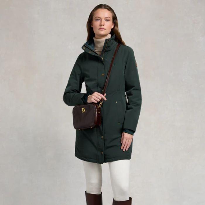 Woman wears a dark green hooded rain coat and cross body bag on a plain background