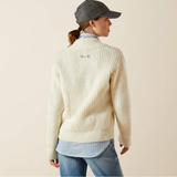 Person wearing a cream ribbed sweater over a light blue shirt with a grey cap on a beige background