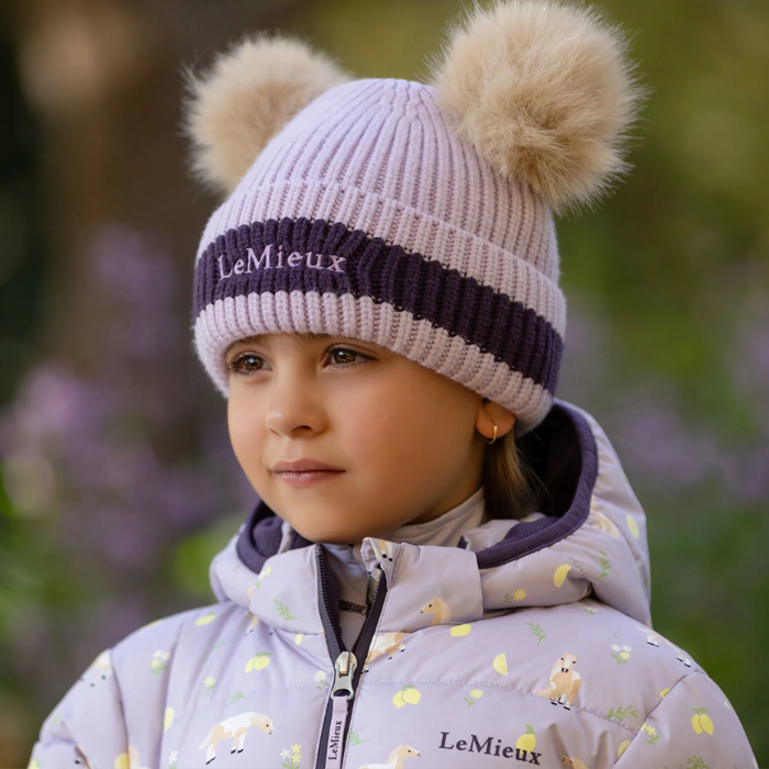 Girl wears a lilac beanie hat and lilac coat in a blurred outdoor setting