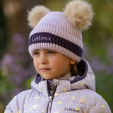 Girl wears a lilac beanie hat and lilac coat in a blurred outdoor setting