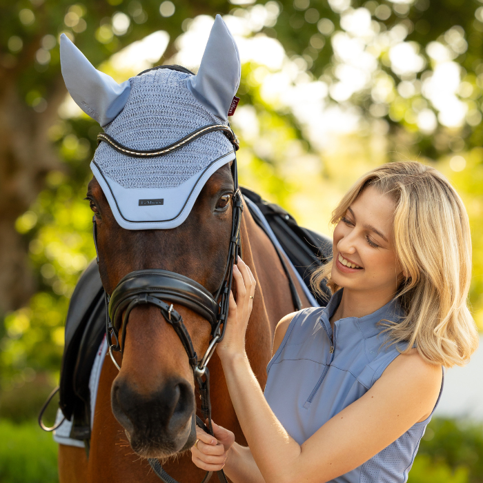 Woman stands with a horse wearing a pale blue fly hood in an outdoor setting