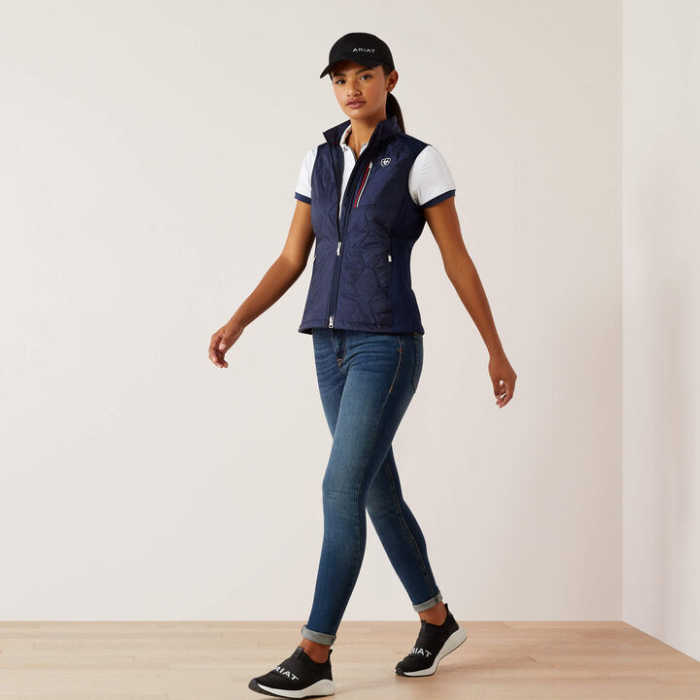 Woman wearing a navy gilet, white shirt, blue jeans, and black cap on a light background