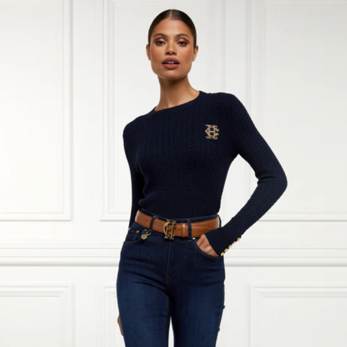 Woman wearing a navy knitted sweater with a logo and jeans against a white background
