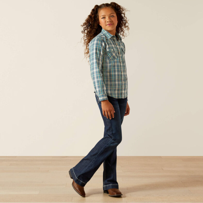 Child wearing a plaid shirt and jeans standing on a wooden floor with a white wall background