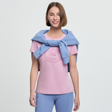 Woman wearing a pink t-shirt with a logo and blue leggings, with a blue sweater over her shoulder on a light grey background.