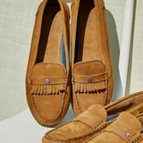 Pair of brown suede loafers with fringe details on a light background