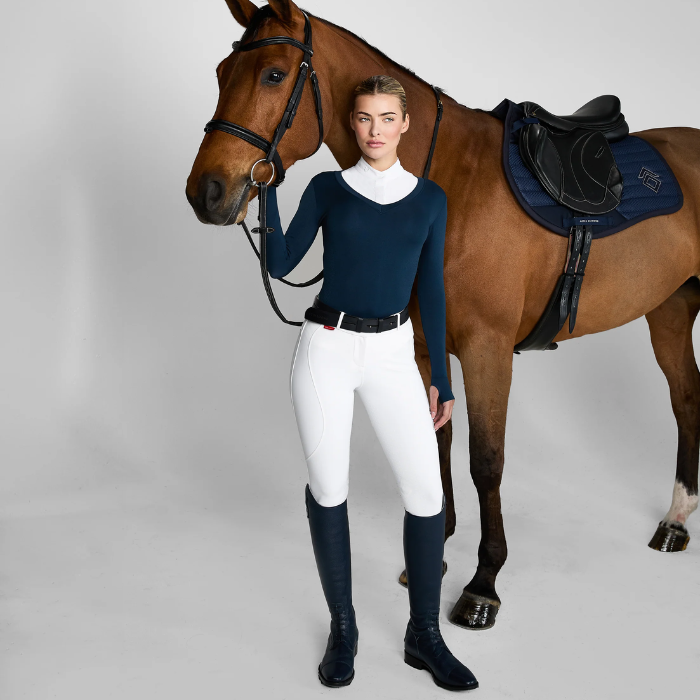 Woman stands with a horse wearing a white shirt, breeches and navy blue sweater on a grey background