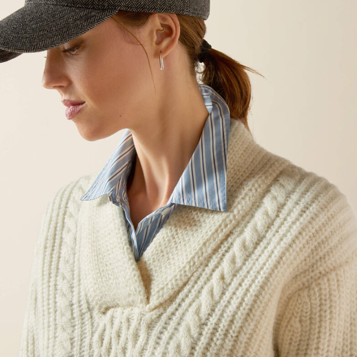 Woman wearing a cream sweater with a striped shirt underneath and a dark cap.