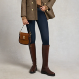 Person wearing a brown coat, blue jeans, and brown knee-high boots, holding a matching brown handbag.