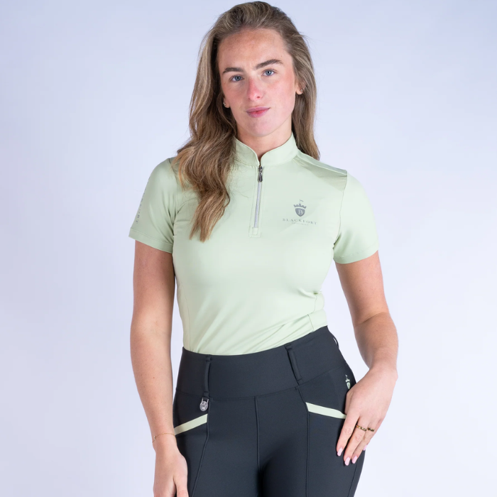 Woman wearing a light green equestrian shirt and black riding leggings on a white background