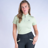 Woman wearing a light green equestrian shirt and black riding leggings on a white background