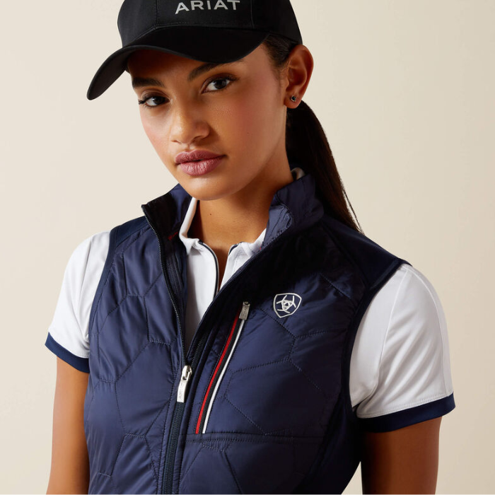 Woman wearing a navy gilet, white shirt, and black cap with 'Ariat' branding on a beige background