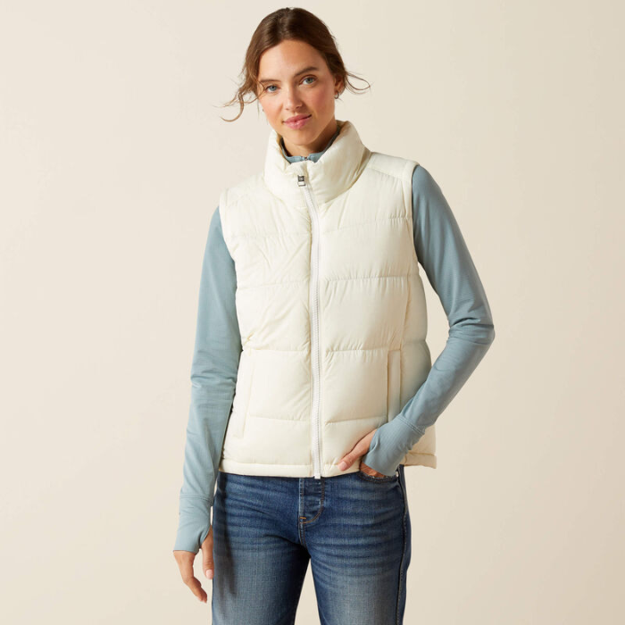 Woman wearing a white puffer gilet over a light blue long-sleeve shirt and jeans on a beige background