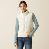 Woman wearing a white puffer gilet over a light blue long-sleeve shirt and jeans on a beige background