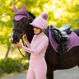 Rider stands holding a horse wearing pink saddle pad and matching ear protectors in a blurred outdoor setting