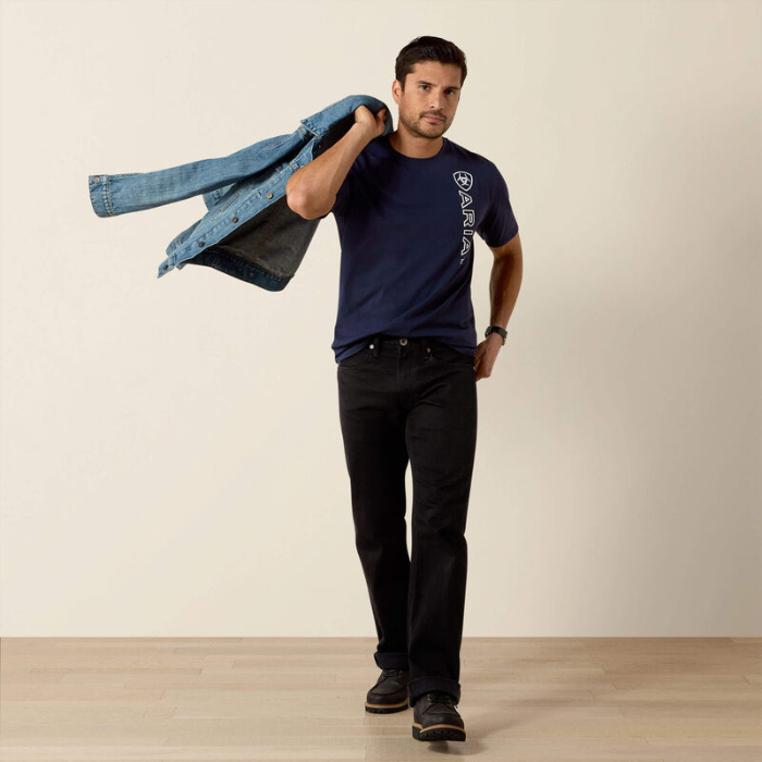 Man wearing a navy blue t-shirt and black jeans, holding a denim jacket against a beige background