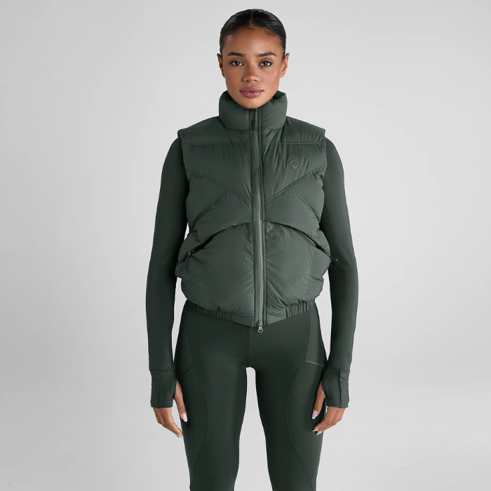 Woman wears a green gilet over an equestrian outfit on a grey background