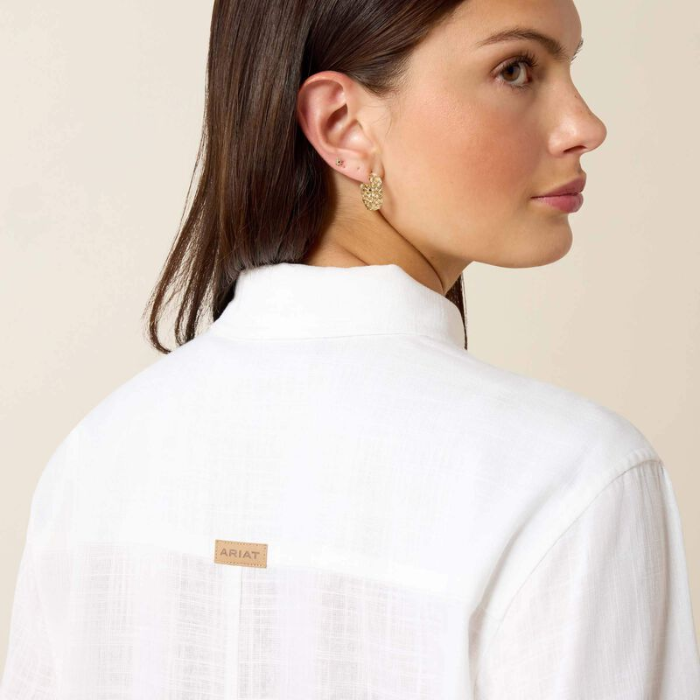 Woman wears a white shirt with ARIAT branding on a beige background