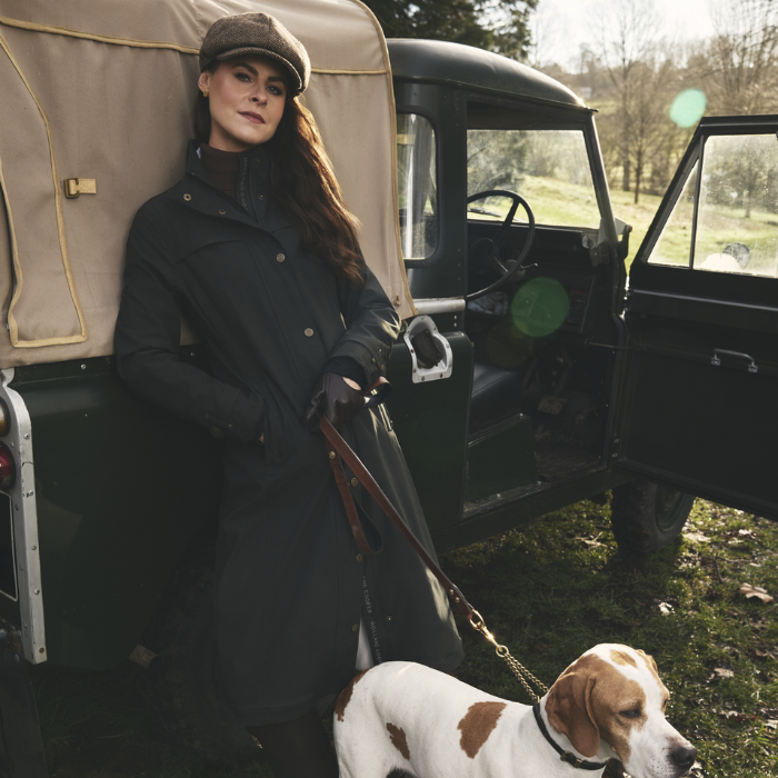 Woman standing next to a vintage vehicle with a dog on a leash in a natural setting