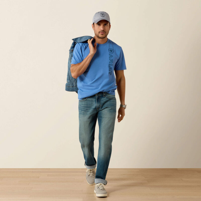 Man wearing a blue t-shirt, jeans, and cap holding a denim jacket against a beige wall.