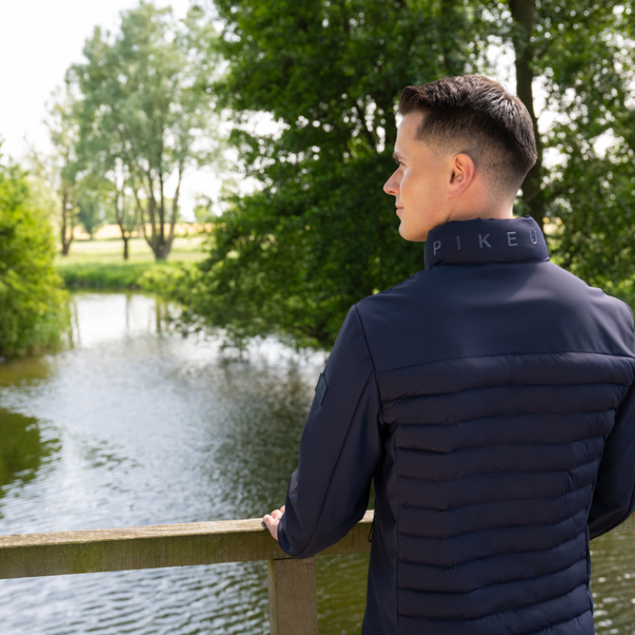 Man wearing a navy blue jacket with 'Pikeur' branding, standing by a body of water with trees in the background.